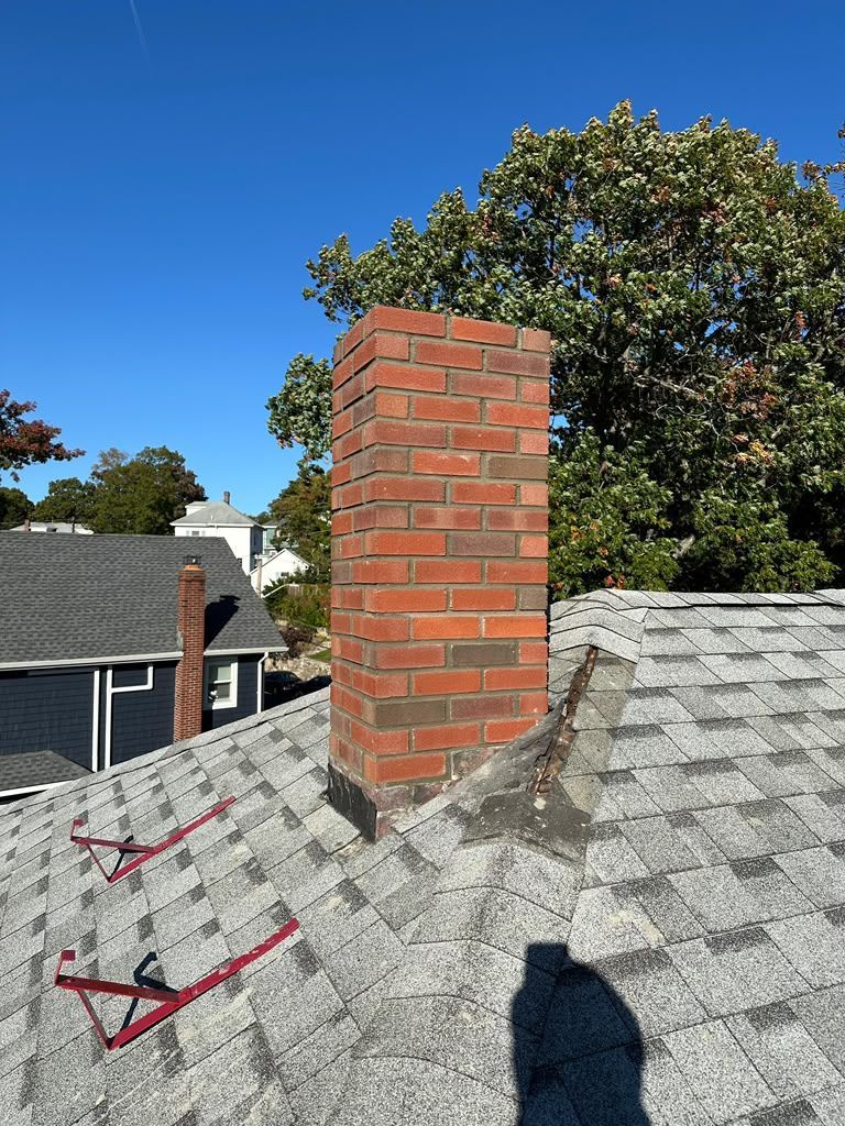 Brick chimney on a shingled roof under a clear blue sky, with trees and neighboring houses in the background.