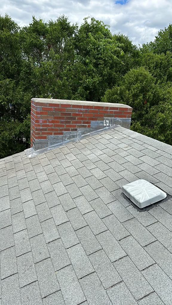 Shingled rooftop with brick chimney and a small vent, surrounded by green trees under a cloudy sky