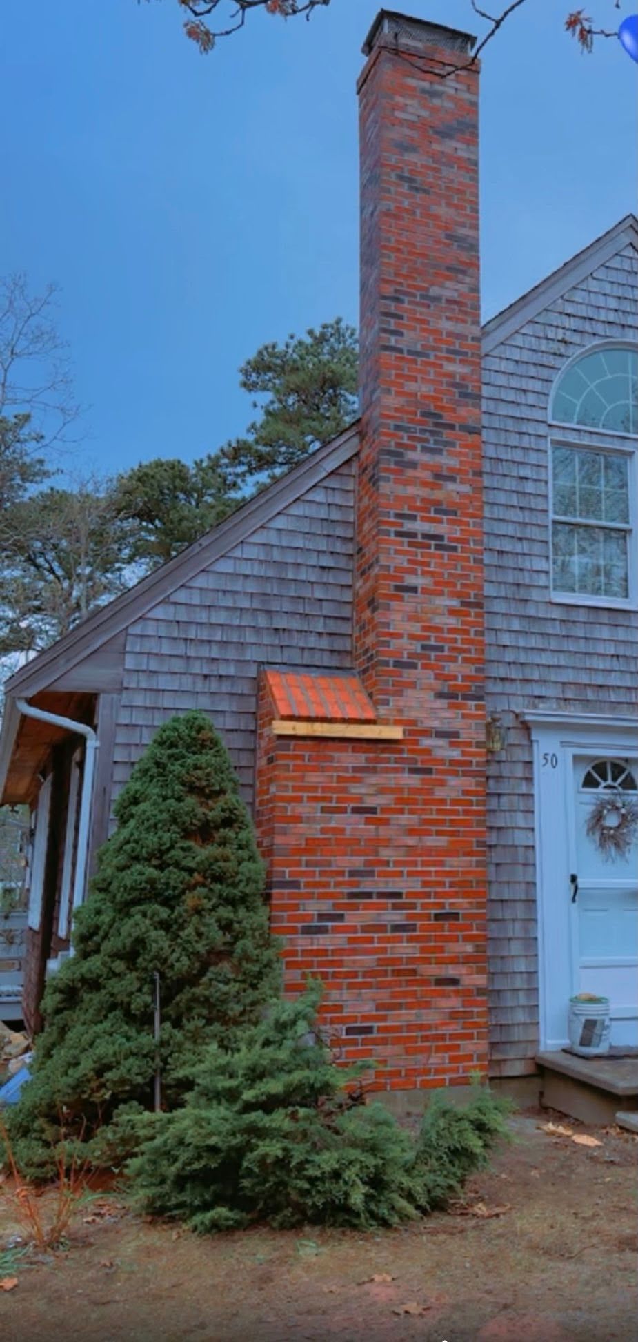 Brick chimney beside a gray house with a white-trimmed window and small evergreen tree in front