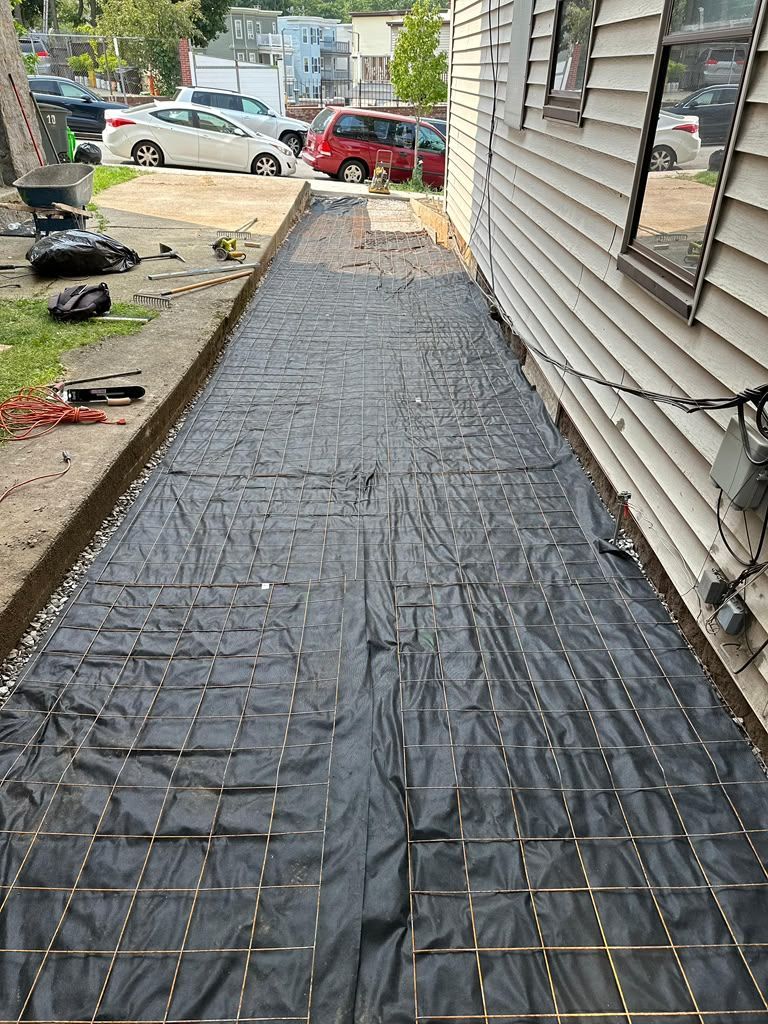 Black landscape fabric covers a narrow side yard beside a house, with driveway and parked cars in the background.