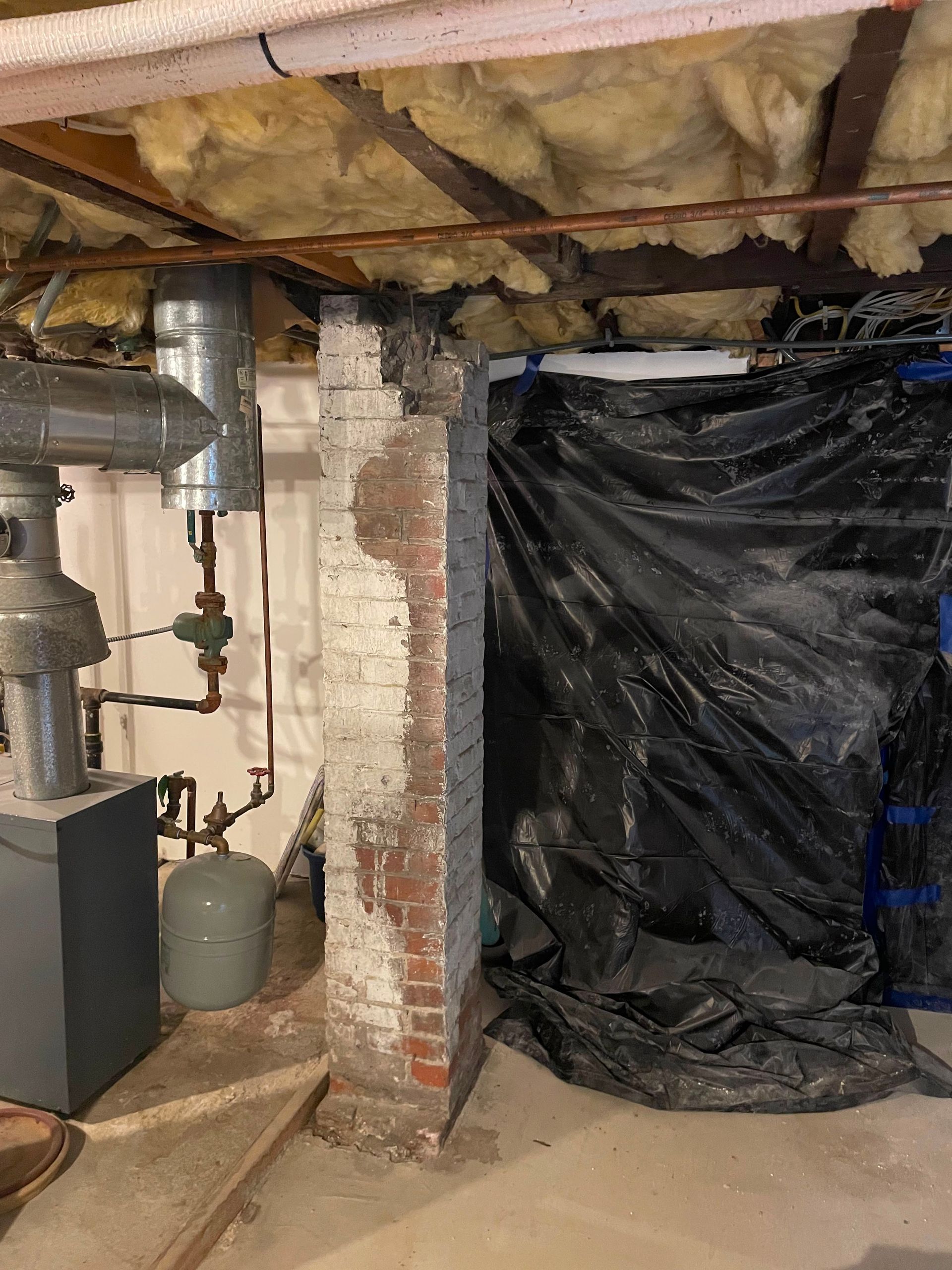 Basement utility area with exposed pipes, insulation, brick wall, and a black plastic-covered opening