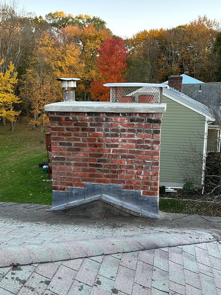 Brick chimney on a rooftop with autumn trees and a house in the background.
