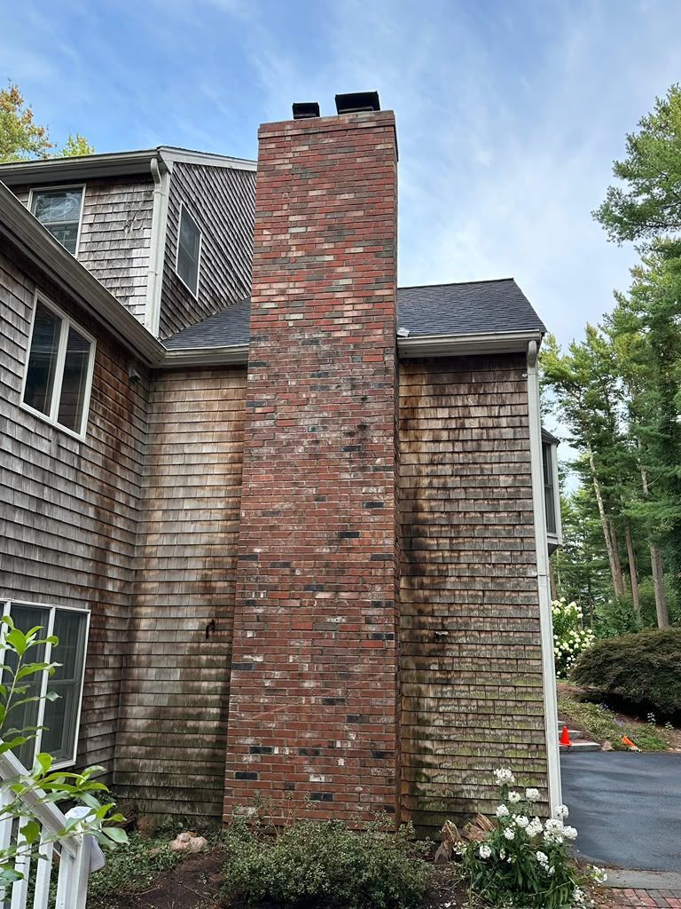 Brick chimney on a shingled house exterior with a chimney cap and surrounding trees.