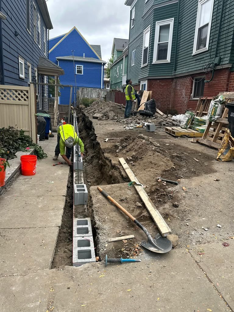 Workers installing a drainage trench along a narrow alley beside houses and a sidewalk