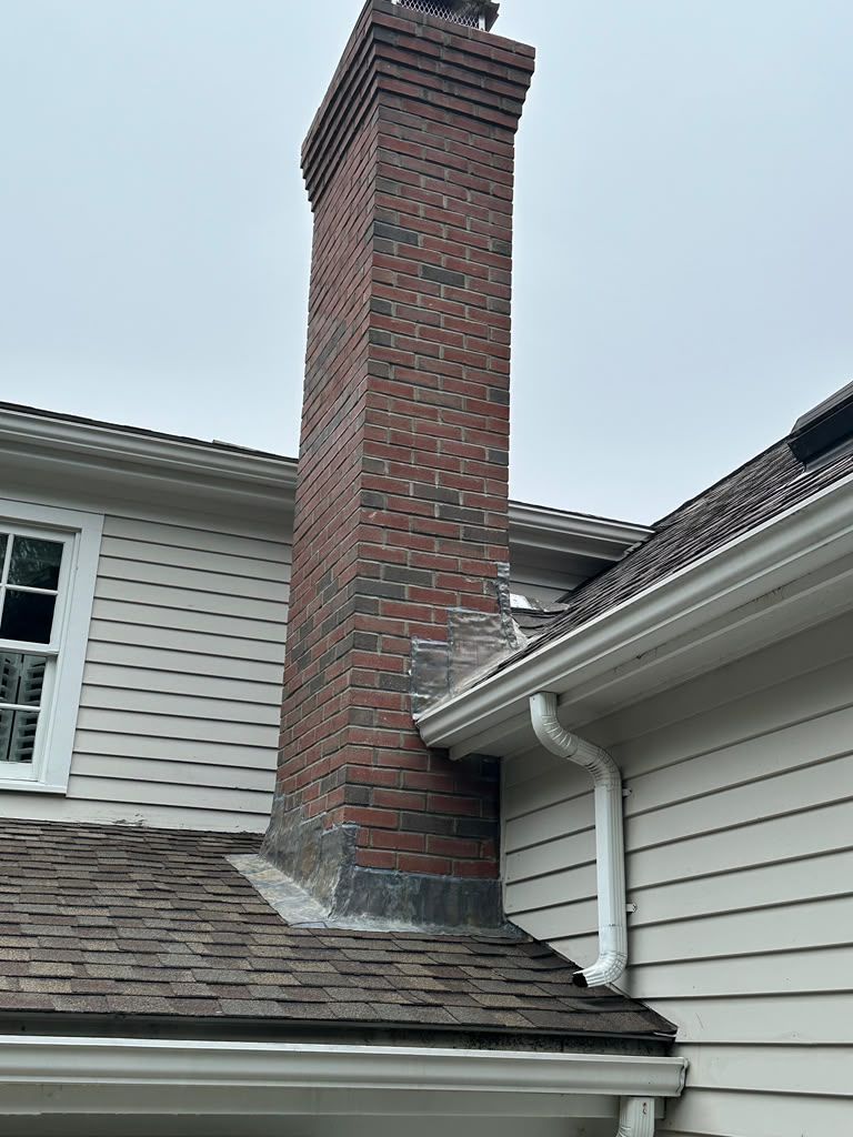 Brick chimney on a gray house with white trim and shingled roof under an overcast sky