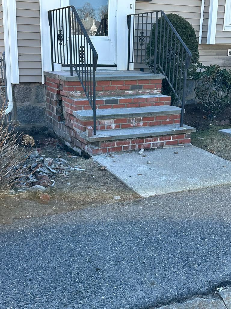 Front steps to a house with brick stairs, black railings, and a concrete landing beside a driveway.