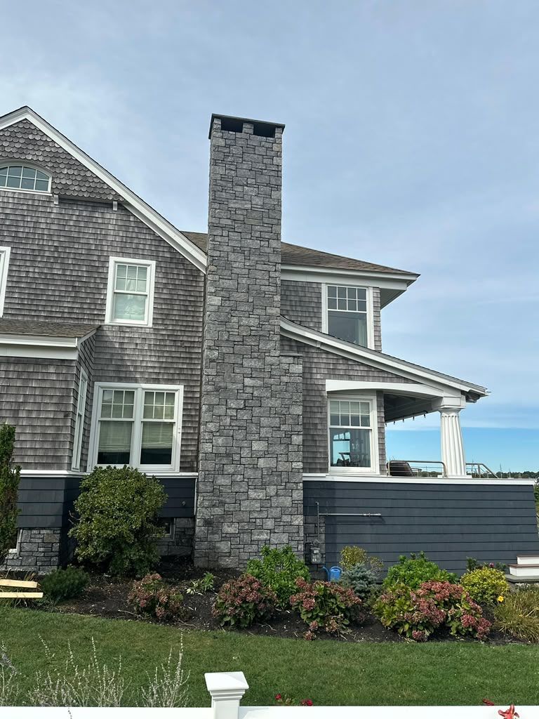 Stone house exterior with tall chimney, gray shingle siding, flower beds, and ocean view under a blue sky