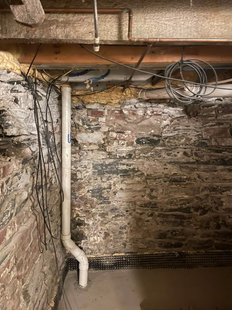 Basement stone wall with exposed pipes, wires, and a damp concrete floor