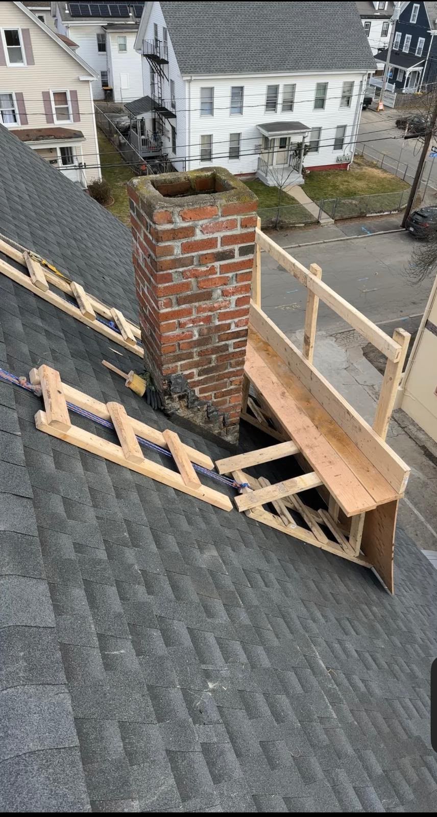 Rooftop repair with wooden framing and a brick chimney beside gray shingles, overlooking houses below