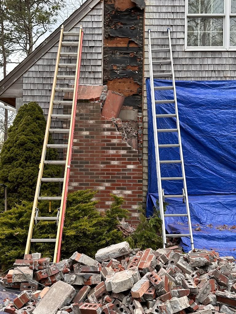 Damaged brick chimney on a house, with debris below and two ladders leaning nearby.