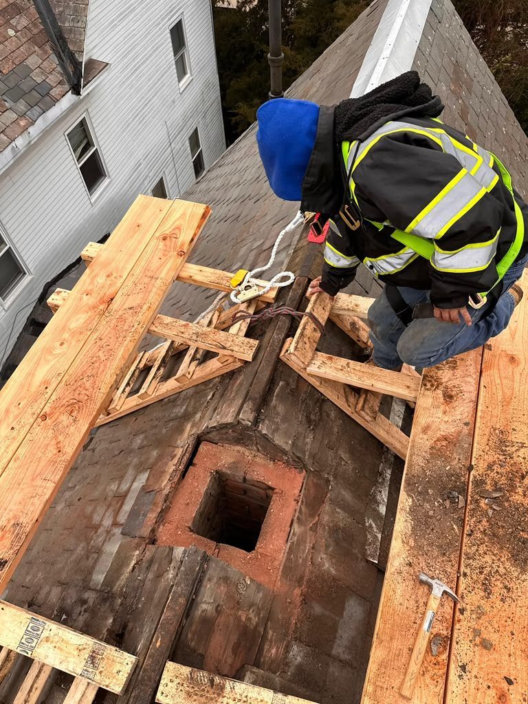 Worker on a roof repairing a damaged section around a square opening, with tools and wood planks nearby.