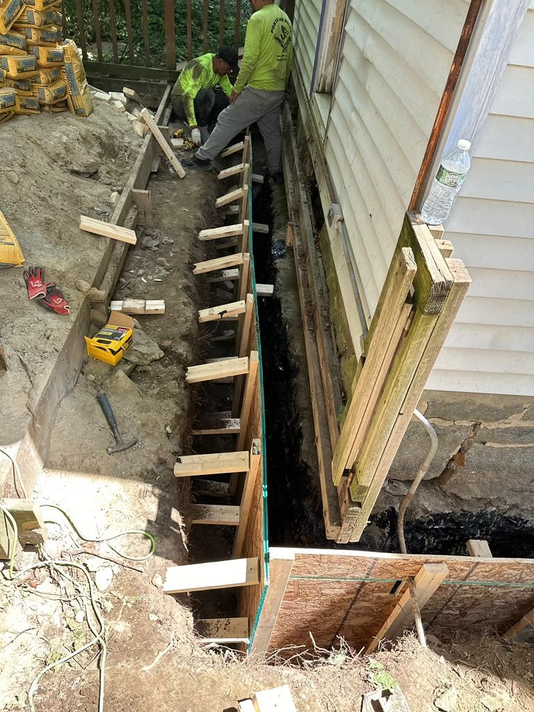 Workers building a concrete foundation beside a house, with wooden forms and a trench