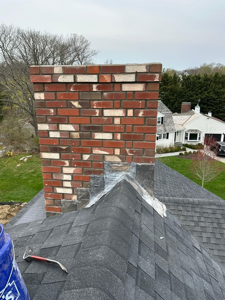 Brick chimney on a shingled roof with white roof flashing, viewed above a residential neighborhood.