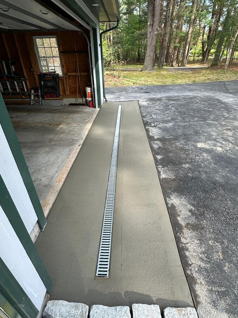Concrete wheelchair ramp with a central drainage channel beside a garage and gravel driveway