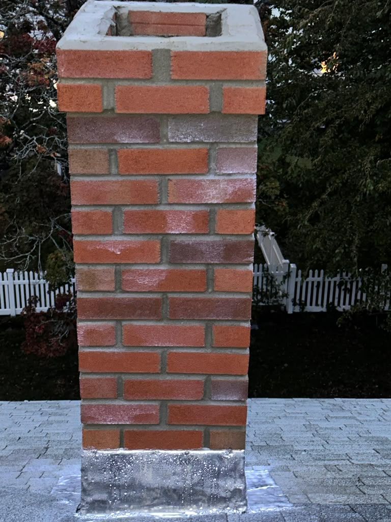 Brick chimney with a concrete cap on a rooftop, set against dark trees and white fence boards.