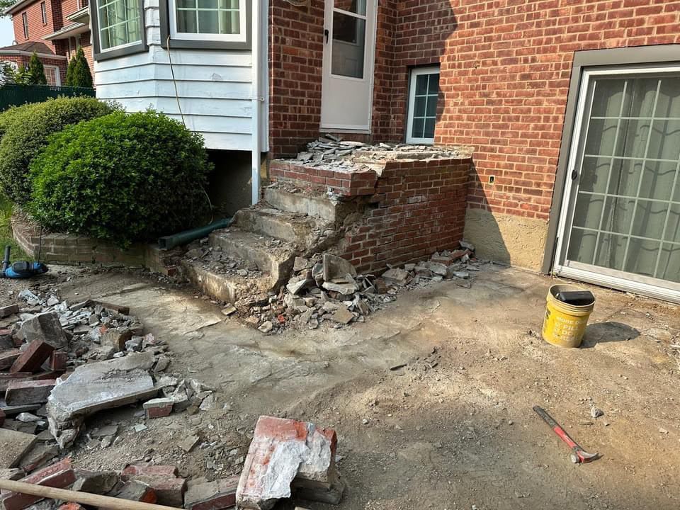 Damaged brick steps and crumbling masonry beside a house, with rubble on the ground and a yellow bucket nearby