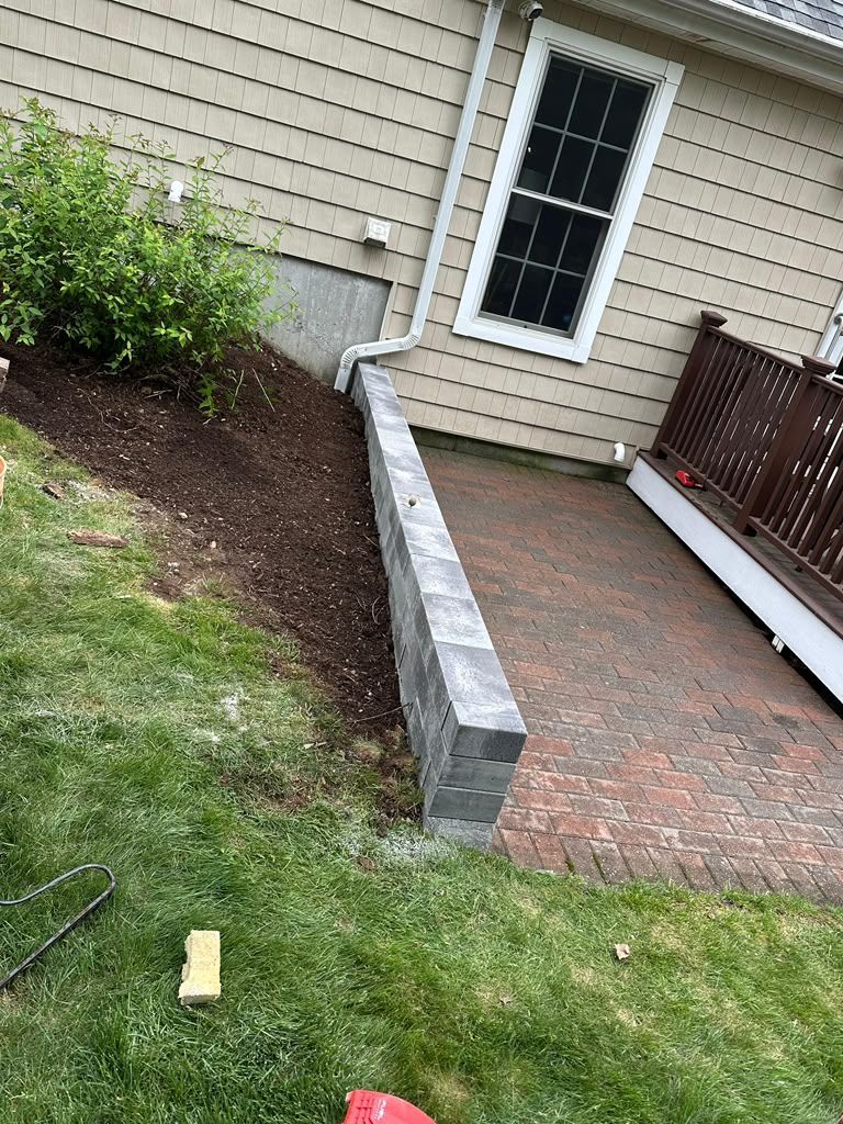 Side yard with brick patio, low cinder block wall, house siding, and a window beside grass and shrubs