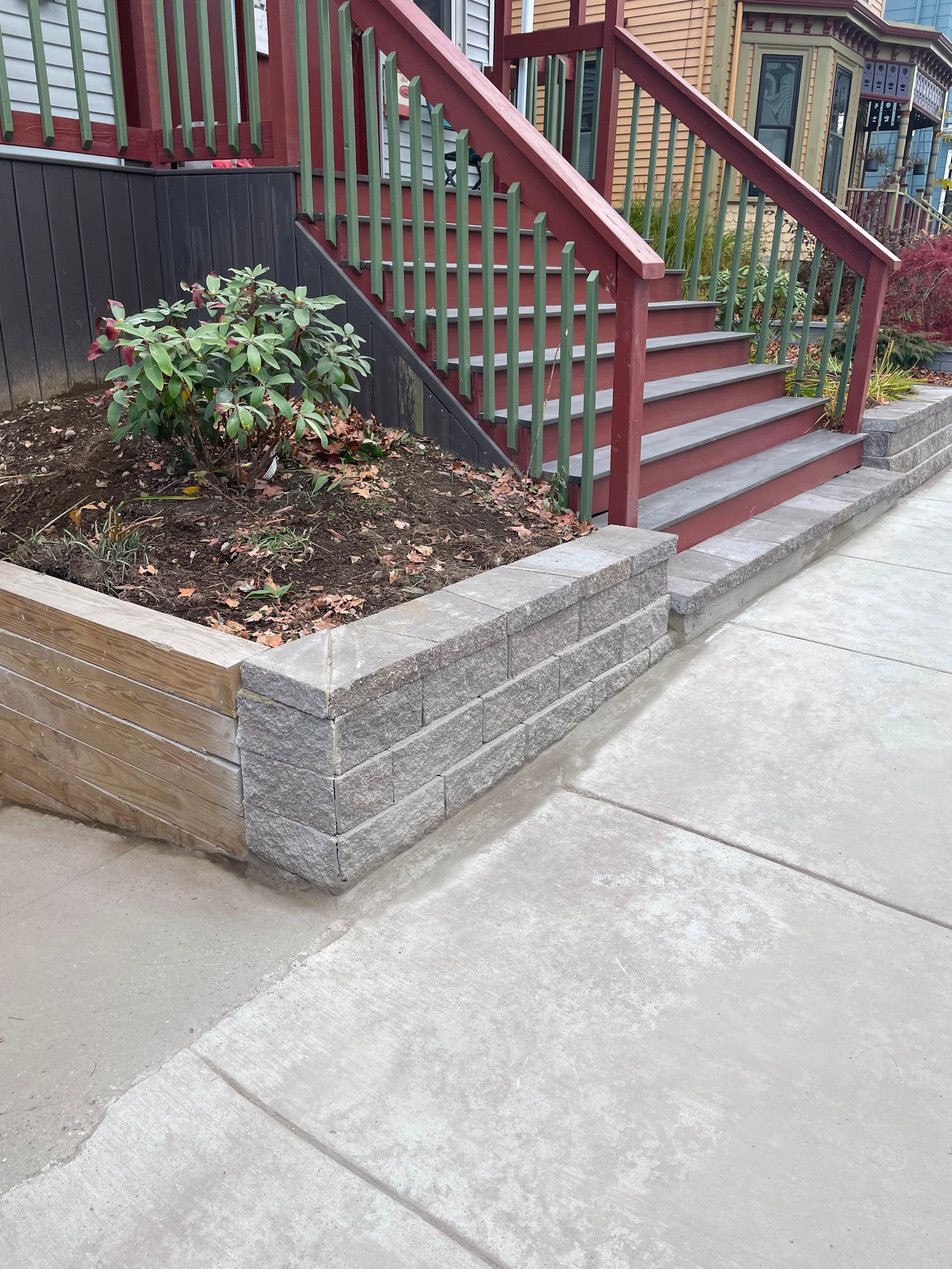 Front steps with red railings beside a small landscaped bed and concrete sidewalk.