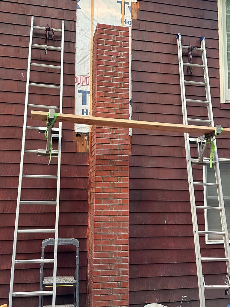 Two ladders braced against a house, supporting a wooden plank beside a brick chimney.