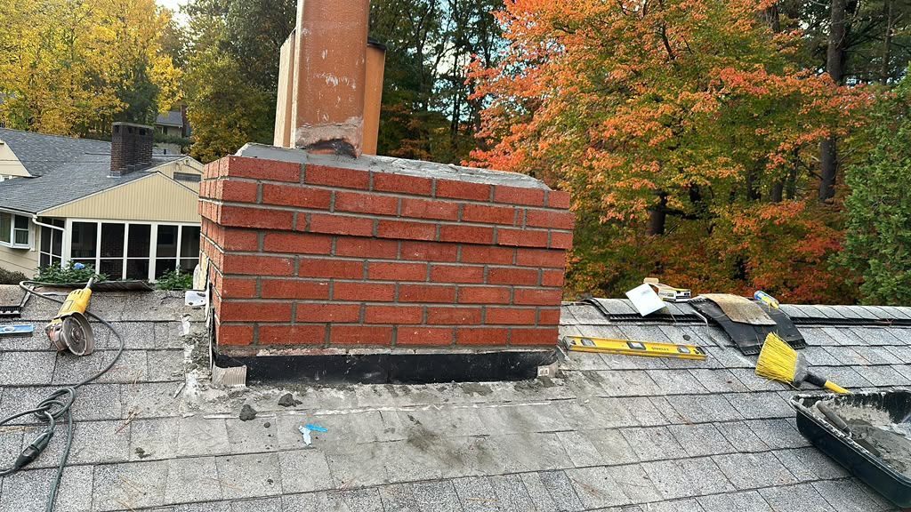 Brick chimney on a shingled roof with autumn trees in the background