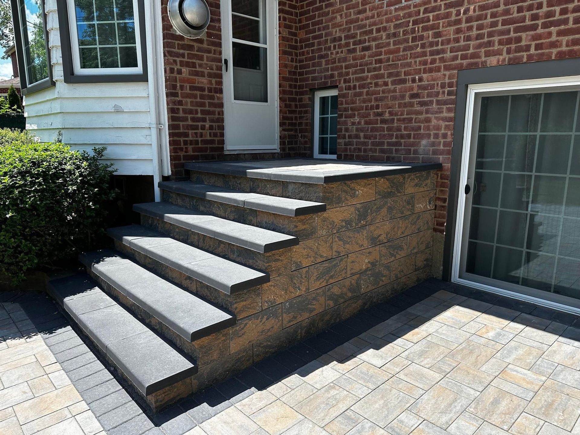 Front steps with white treads leading to a brick house entrance, beside a large window and paved patio.