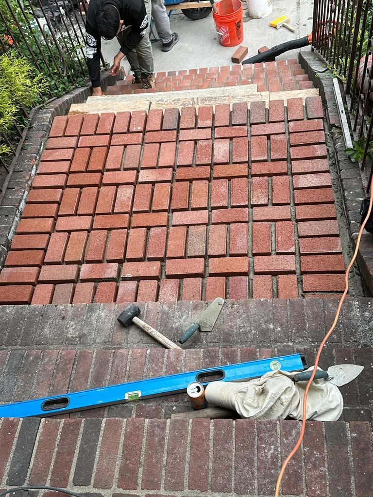 Brick walkway under construction with level, tools, and workers in the background