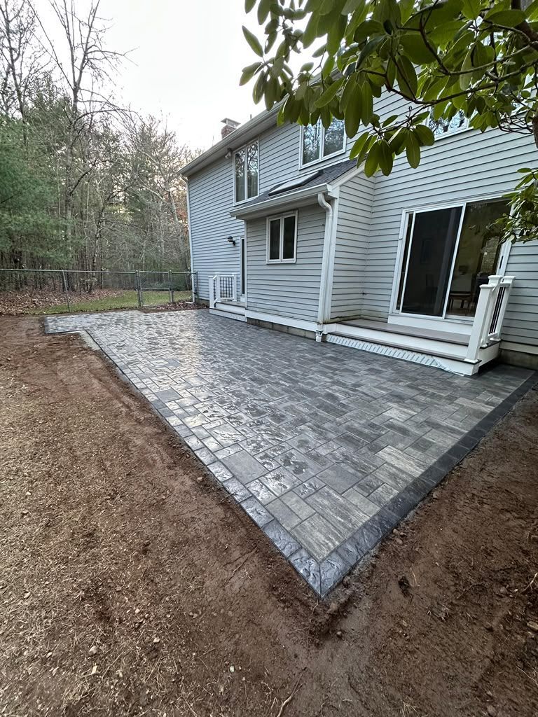 Stone-paver patio beside a white house with steps and sliding glass doors, bordered by trees and mulch.