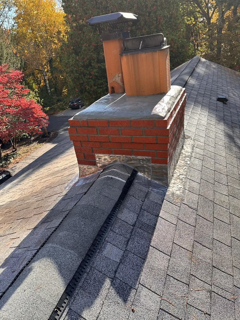 Rooftop view of a brick chimney with a metal cap and gray shingles in autumn light