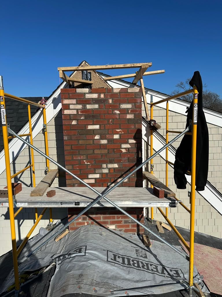 Scaffolded brick chimney under construction against a blue sky, with a tarp-covered base.