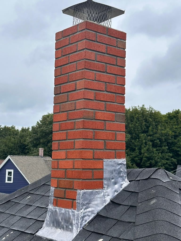 Red brick chimney with metal cap and flashing on a shingled rooftop against cloudy sky.