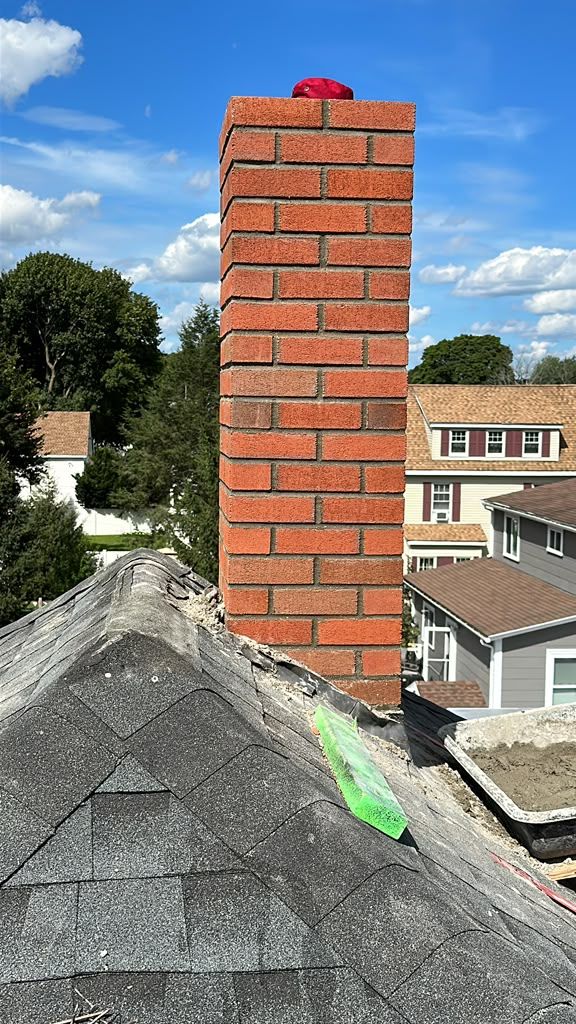 Brick chimney on a shingled rooftop with a red cap against a cloudy sky.