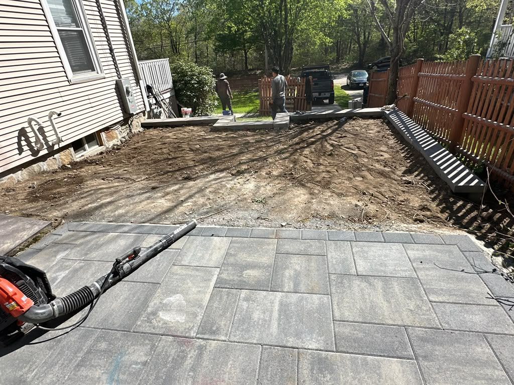 Backyard patio under construction with paver stones and a dirt work area, bordered by a fence and house