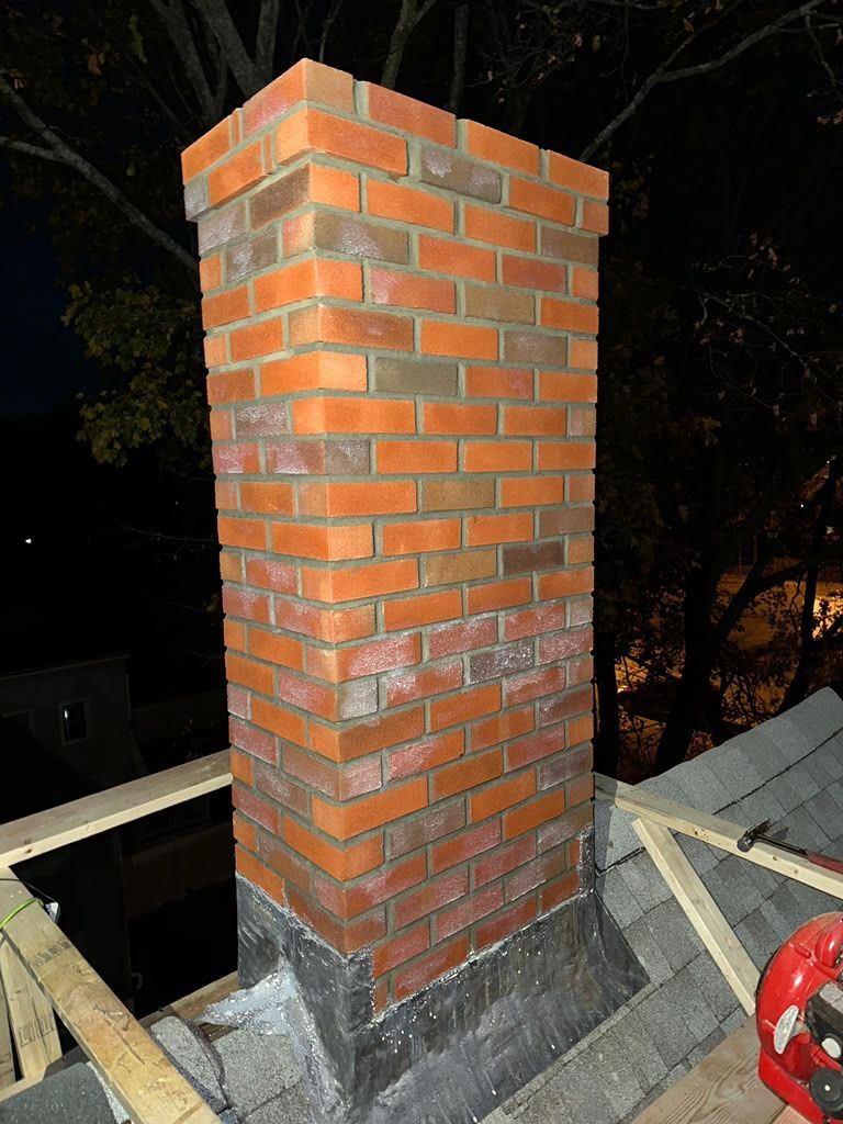 Brick chimney on a rooftop at night, with unfinished concrete base and construction materials nearby
