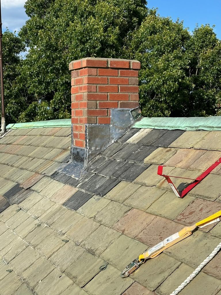 Rooftop chimney with brick and metal flashing on a shingled roof, with tools and trees in the background