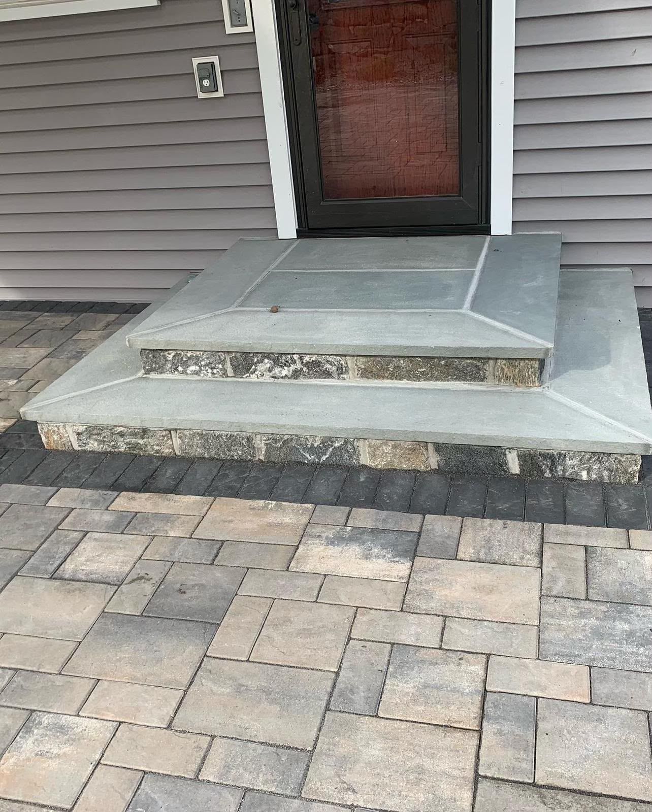 Front entry with concrete steps and platform, brick paver walkway, and a closed door on gray siding.