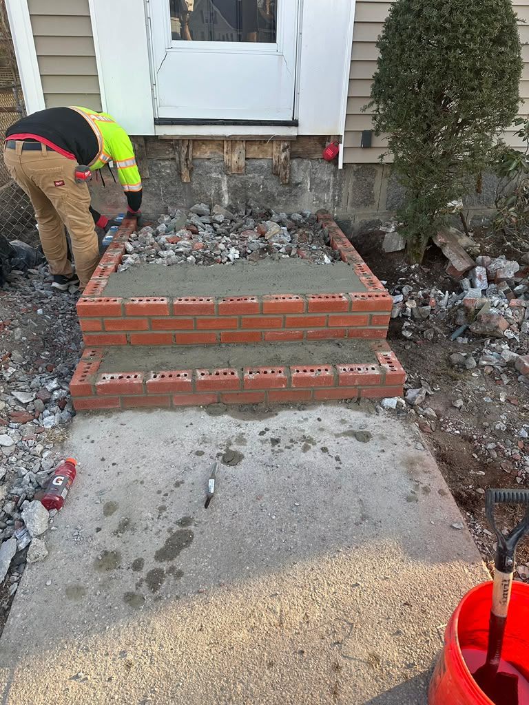 Worker rebuilding red brick front steps beside a white door, with rubble and tools on a walkway.