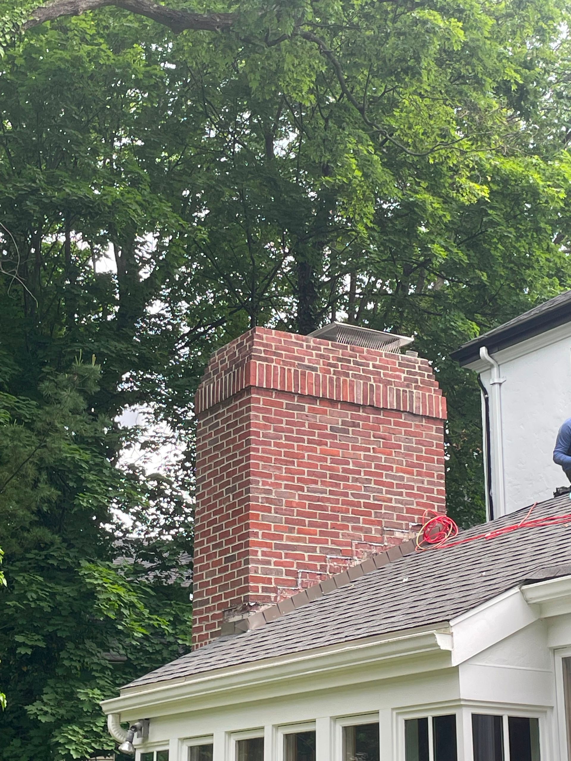 Brick chimney on a shingled roof beside a white house, with tall green trees in the background.