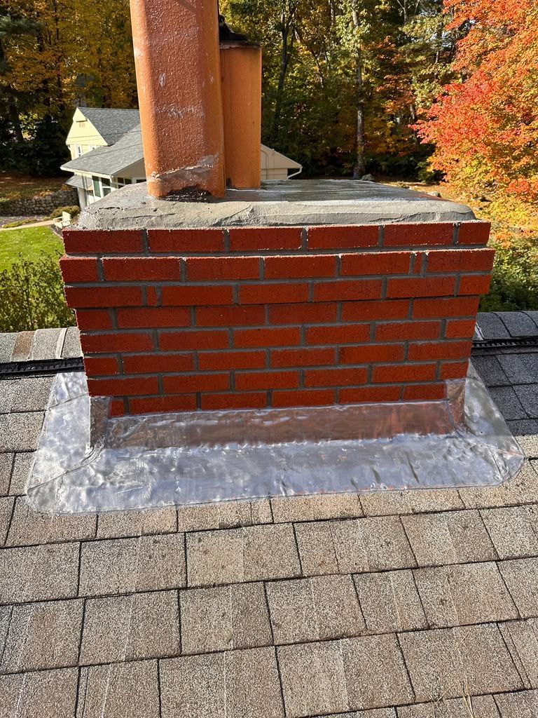 Red brick chimney base on a gray roof with autumn trees in the background