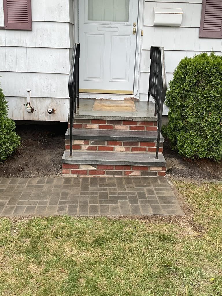 Front steps with brick walkway leading to a white house door, bordered by black handrails and shrubs.