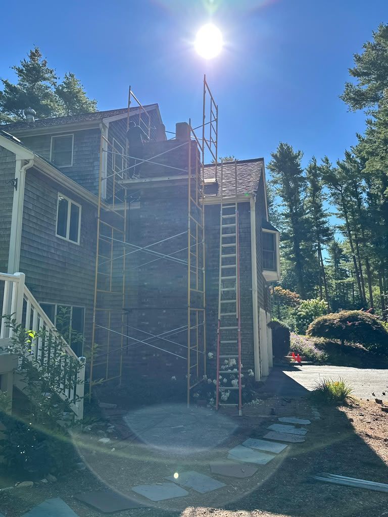 Brick house exterior with scaffolding and ladder in bright sunlight, surrounded by trees