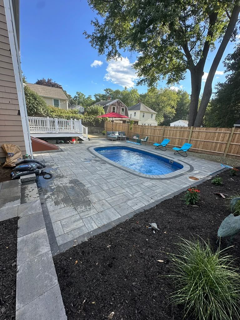 Backyard with a small oval pool, gray patio, blue lounge chairs, and trees under a clear sky