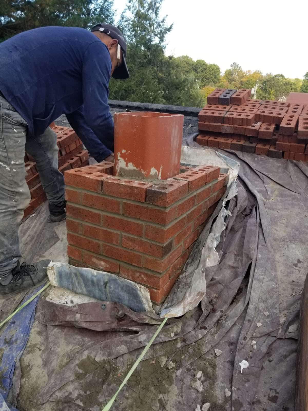 Worker building a brick chimney on a rooftop, with stacks of bricks and tarp-covered surface nearby.