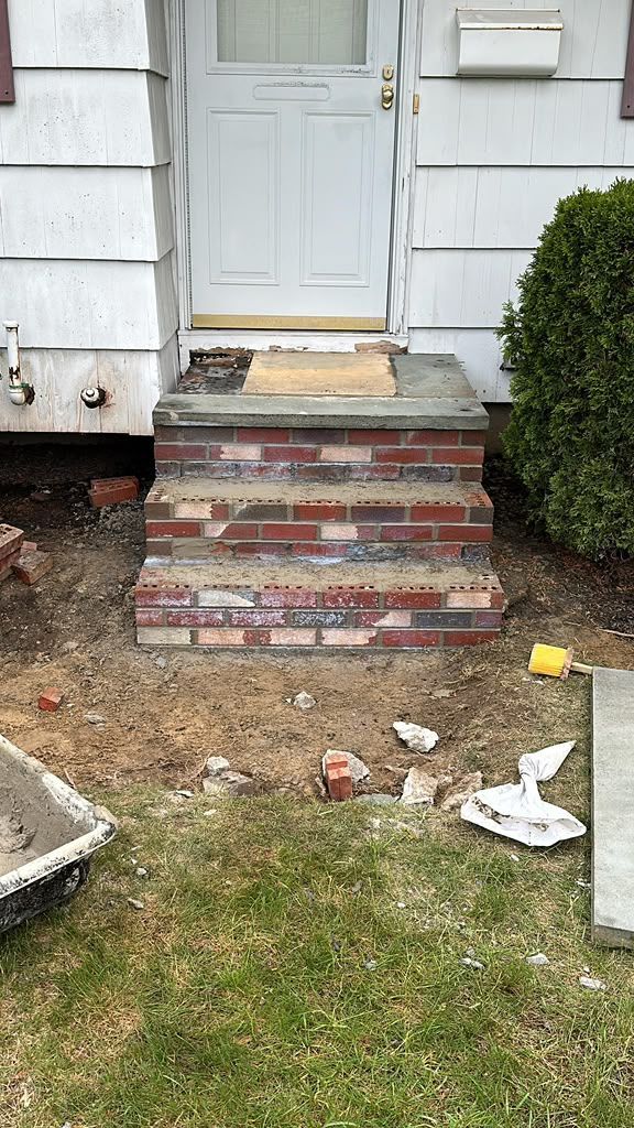 Front porch with brick steps, white door, and a small shrub beside a patchy yard with litter.