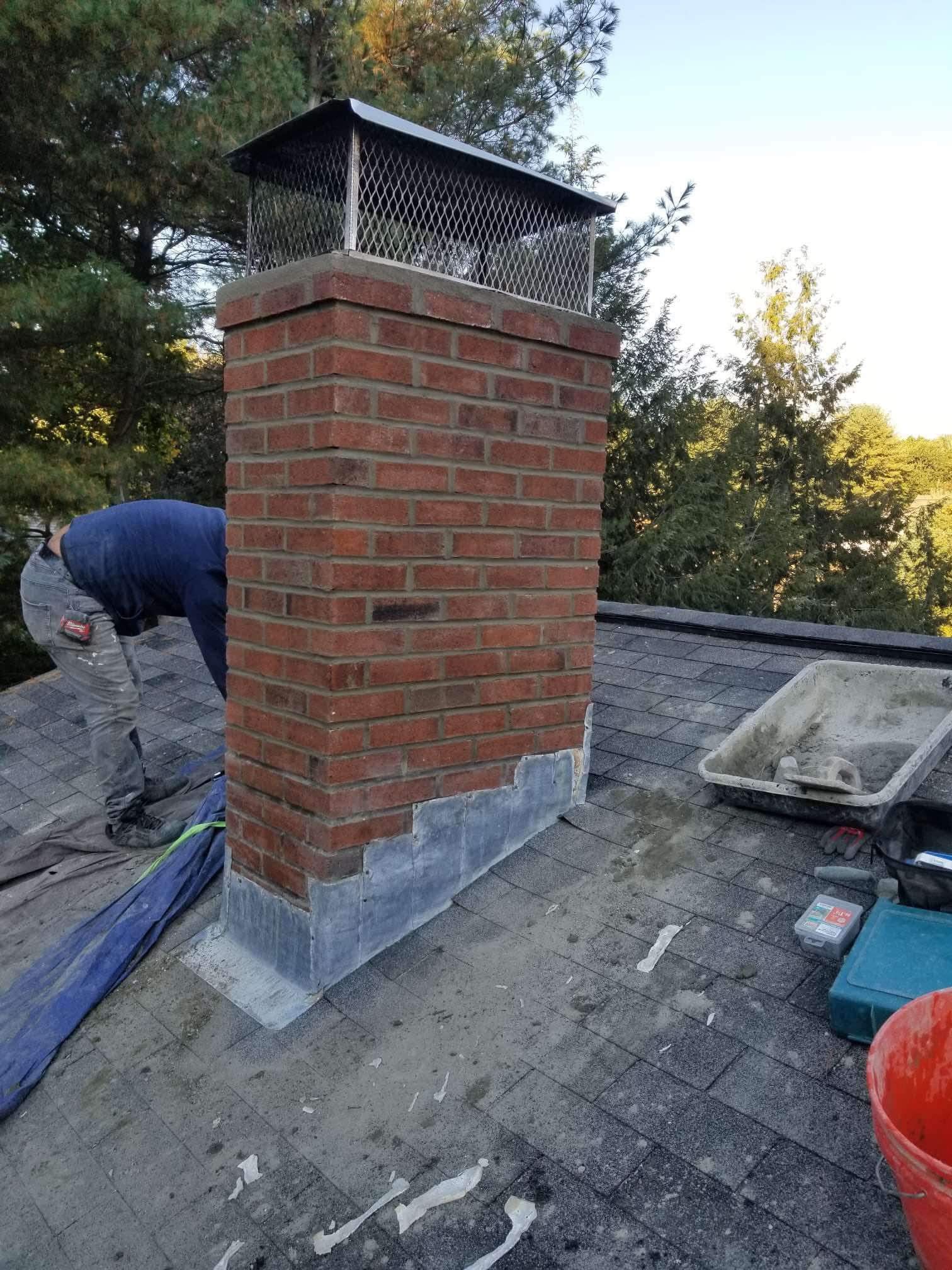 Brick chimney under construction on a shingled roof, with a worker beside it and trees in the background.