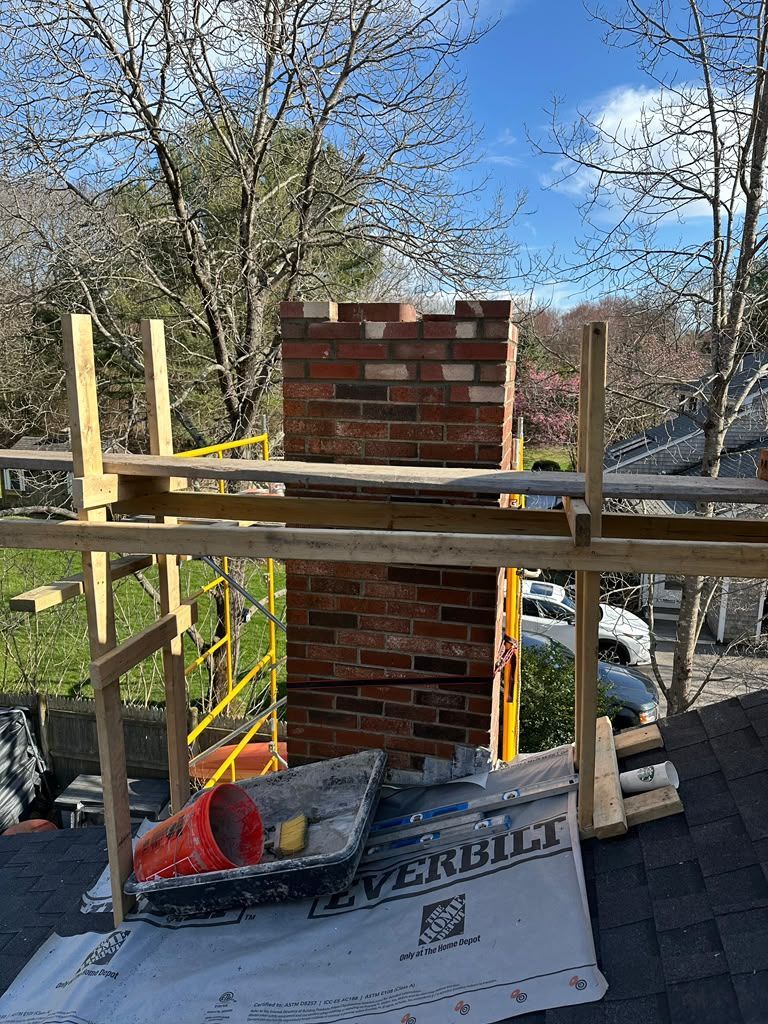 Brick chimney under construction on a scaffolded roof, with lumber, tools, and winter trees in the background