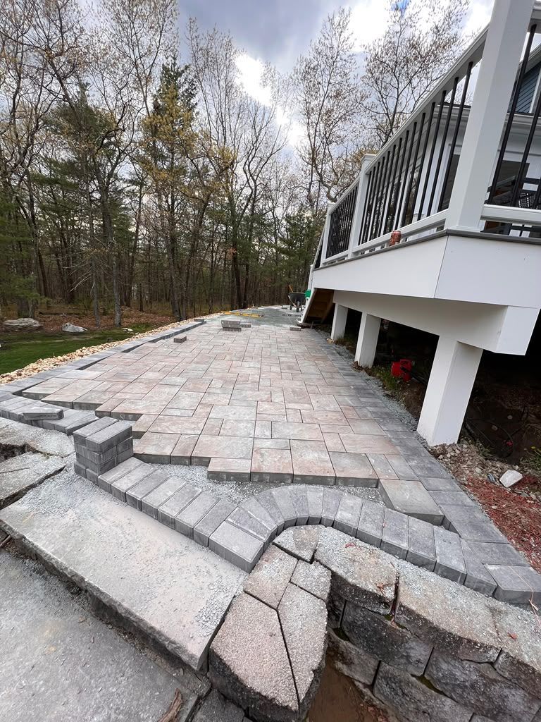 Uneven stone patio beside a house deck, with sunken pavers and wooded background.