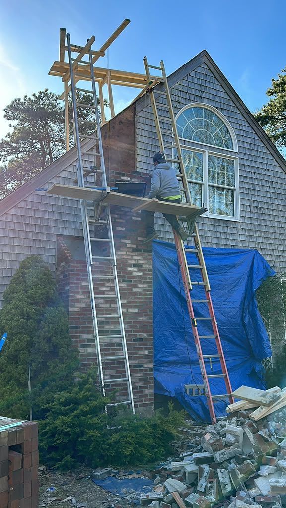 House exterior under renovation with scaffolding, ladders, blue tarp, and stone pile in front