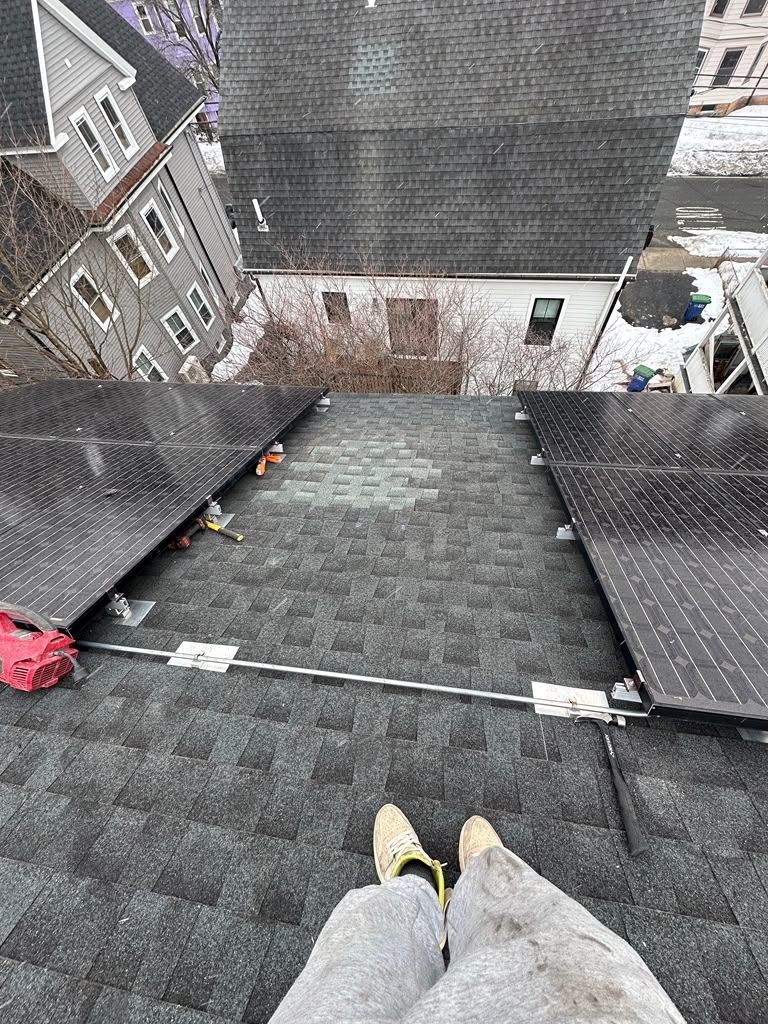 Rooftop view from a person’s feet, showing gray shingles, neighboring buildings, and a red bag on the left edge
