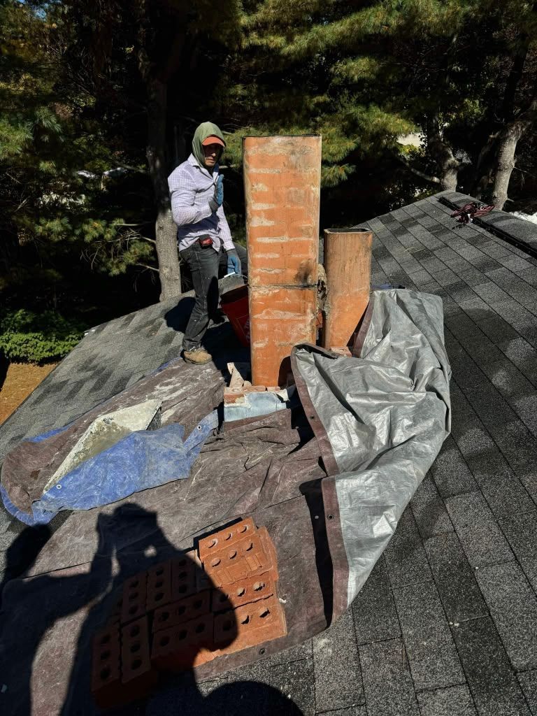 Worker on a shingled roof beside a brick chimney, with tarps and roofing materials scattered around.