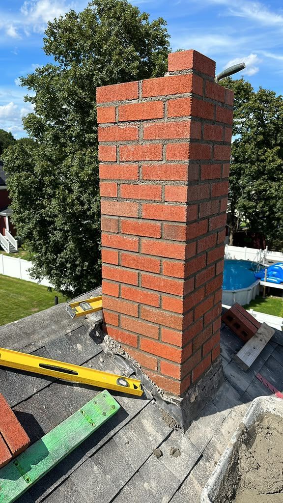Brick chimney stack under construction on a rooftop, with tools and a blue sky in the background